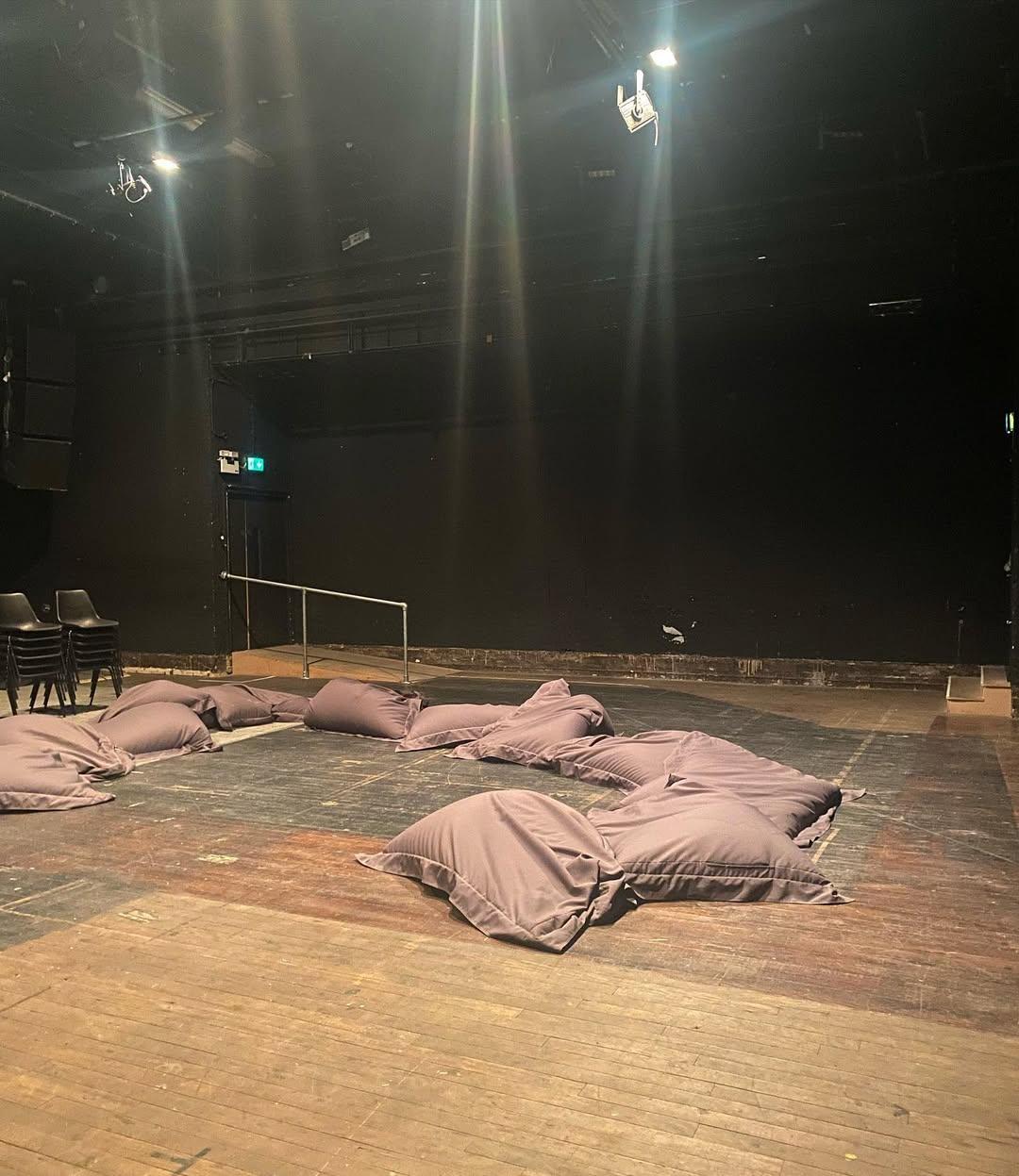 The ICA stage prepared for an event: dusty mauve beanbags scattered across a wooden floor beneath spotlights, stacked black chairs at the edge — a space waiting to be activated