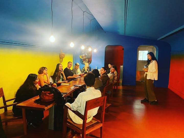 A group seated around a long wooden table inside a room painted in bold blocks of yellow, blue, red, and orange — bare bulbs hanging above, immersive and engaged