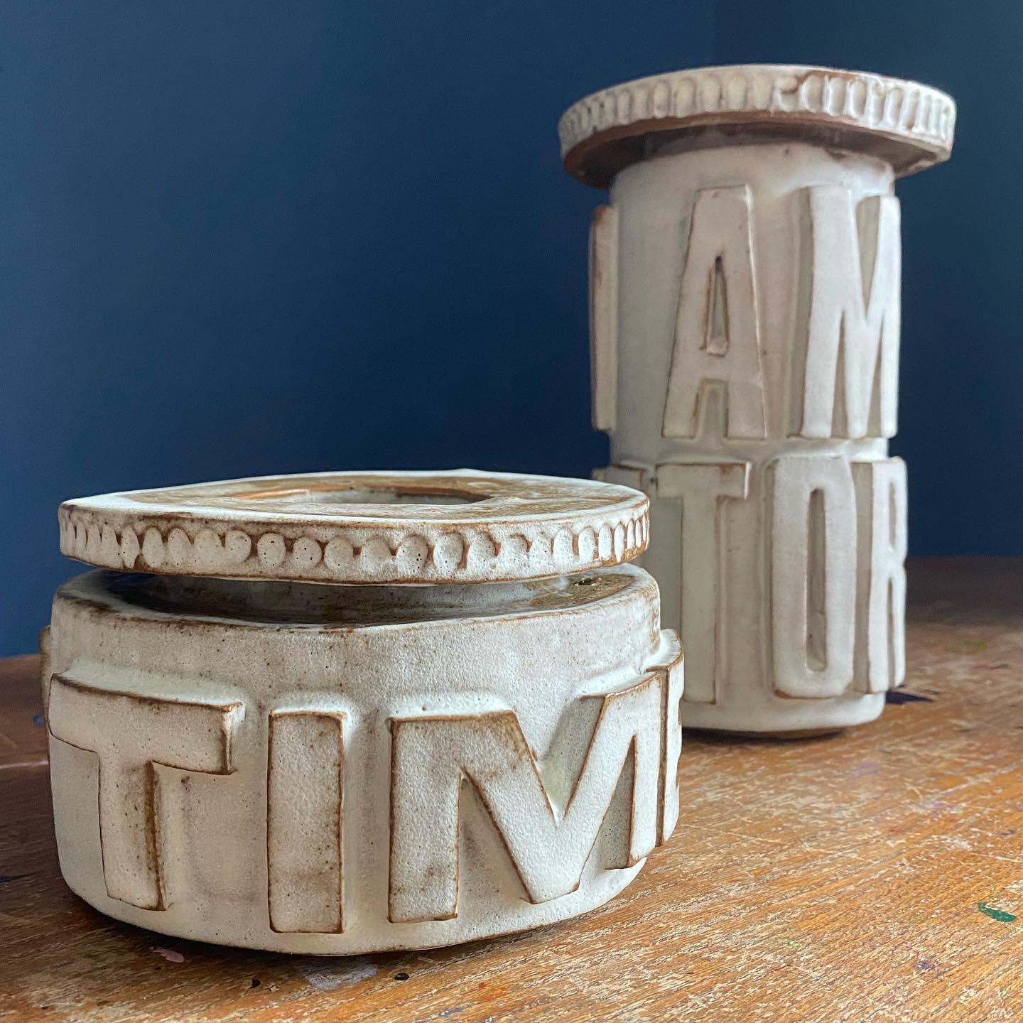 A pair of cream stoneware vessels with raised letters — the squat drum reads TIME, the taller column reads VICTORY — amber glaze pooling in the carved recesses, set against a deep teal wall