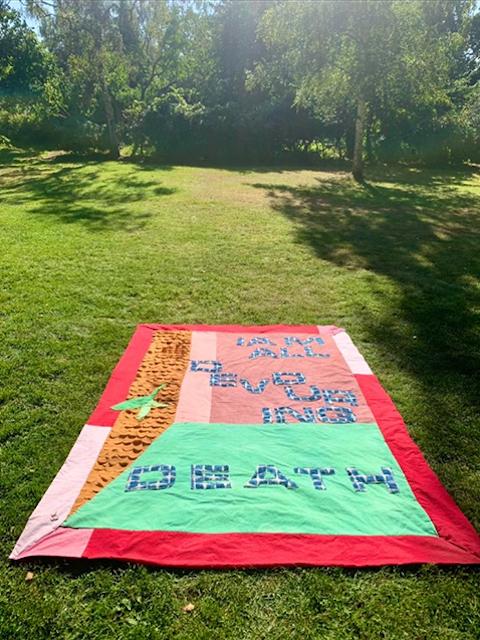 A quilt stretched on a lawn bearing the words ALL DEVOURING DEATH in blue-and-white mosaic-patterned letters, framed by vivid coral borders and a strip of amber — death rendered in the bright palette of the living