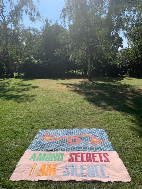 A hand-dyed quilt laid on sunlit grass, an orange gecko resting on blue-and-white geometric fabric, the words AMONG SECRETS I AM SILENCE spelled in bright multicoloured letters across the lower border