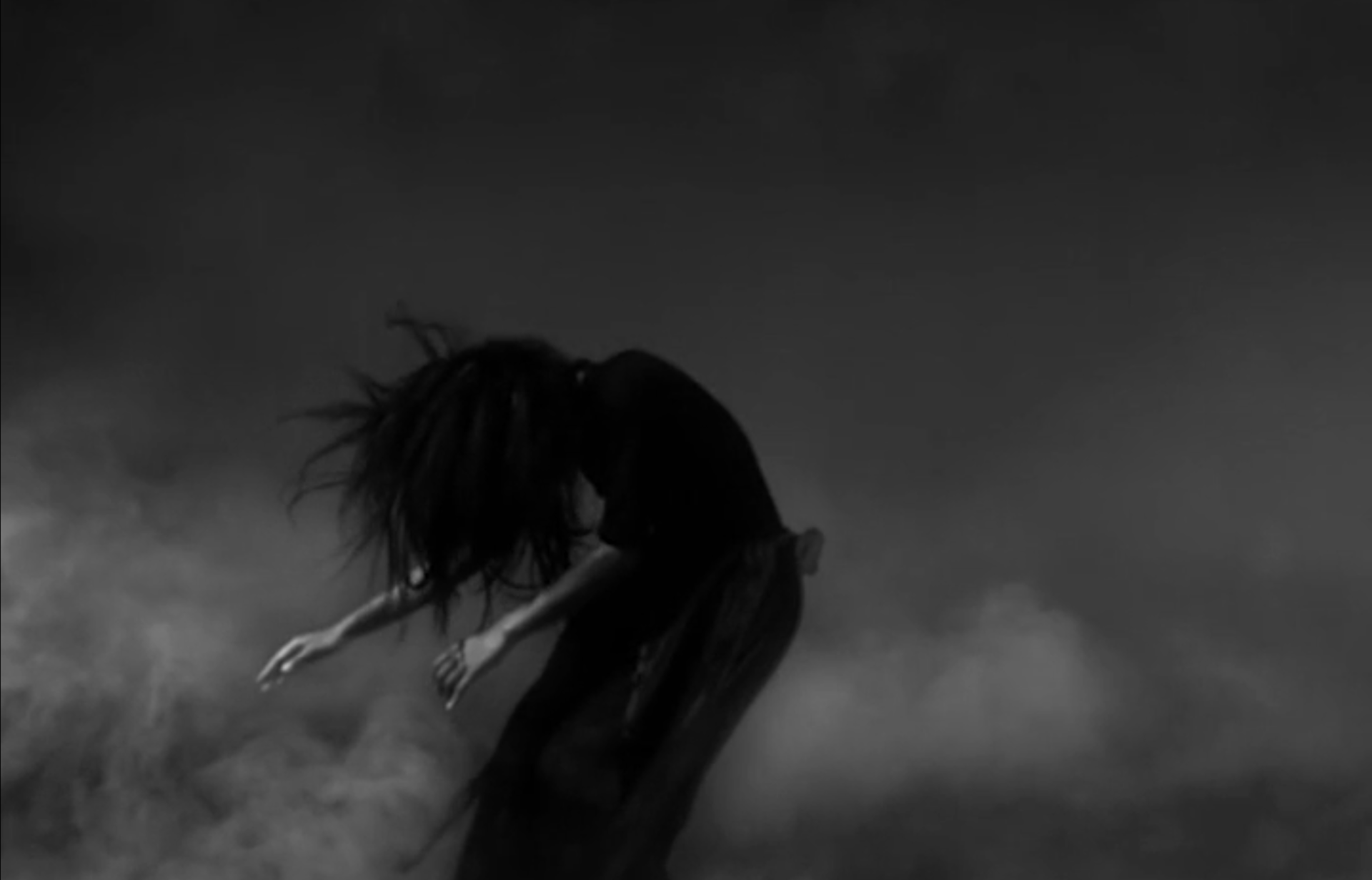A figure hunched in black-and-white, hair wild, arms outstretched, emerging from billowing smoke — a still of anguished transformation, the body as site of resistance