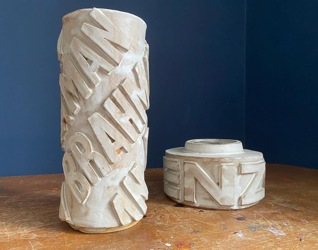 Two stoneware vessels: a tall, slightly leaning cylinder spiralled with the letters BRAHMAN beside a squat drum bearing BENZOS — the transcendent and the sedated, paired in cream clay against navy blue
