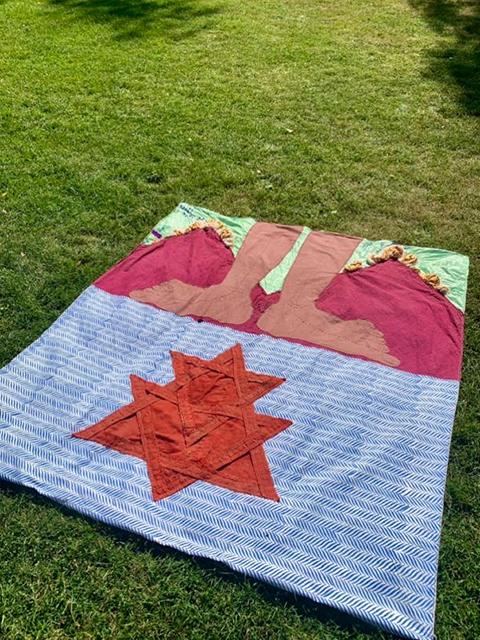 A textile work on green grass: deep magenta and brown body-like forms emerge in the upper half, while a rust-orange star blazes against blue-and-white chevron fabric below — an evocation of feminine power rising from patterned depths