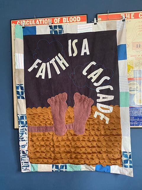 A wall-hung textile with dusty pink feet suspended against a dark ground, the words FAITH IS A CASCADE above, and a golden cascade of overlapping petal-like fabric pieces flowing downward — hung between vintage anatomy charts on a blue wall