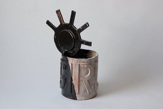 A rough, patchy-glazed ceramic cylinder topped with a dramatic glossy black sun-like form, its spokes radiating outward — the word GIRLS raised on the vessel below, punk energy contained in clay