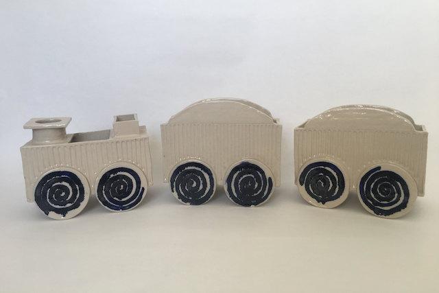 A ceramic toy train of three carriages in cream stoneware, each set on wheels decorated with dark blue spirals — a childhood object rendered in clay, poised between nostalgia and transformation