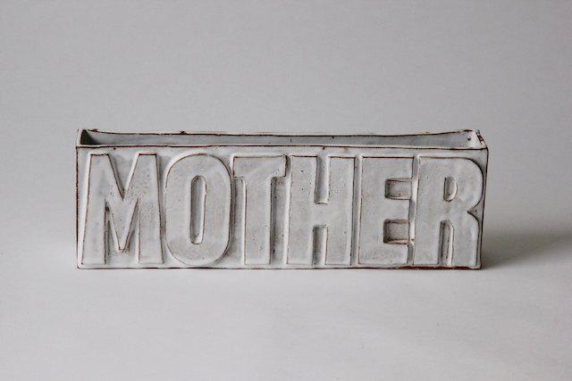 A slab-built ceramic rectangle with the word MOTHER raised in bold relief across its face, white glaze breaking to terracotta at the sharp edges — monumental and tender, like an inscription between gravestone and love letter