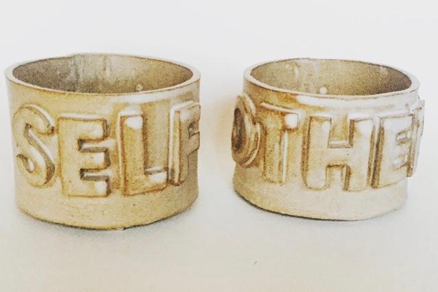 Two small ceramic cylinders the colour of old parchment, one bearing SELF, the other OTHERS — sitting side by side like a philosophical sentence that refuses to close