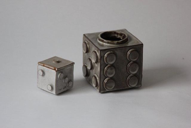 Two ceramic dice — one small and pale, one large in dark gunmetal glaze — their circular pips pressed into the clay, the larger one open at the top as a vessel for flowers or chance